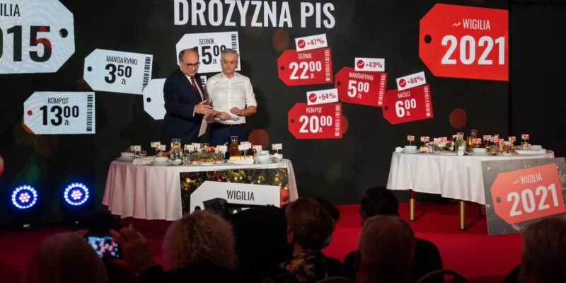 Dwóch mężczyzn w garniturze i białej koszuli stoi przy okrągłym stole zastawionym potrawami podczas wydarzenia Nowej Lewicy „Ulga noworoczna”, z tłem z cenami i napisem Wigilia 2021.