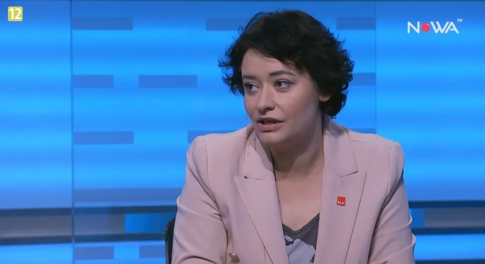 Kobieta w jasnoróżowej marynarce siedzi i rozmawia w studiu telewizyjnym na niebieskim tle z widocznym logotypem stacji.