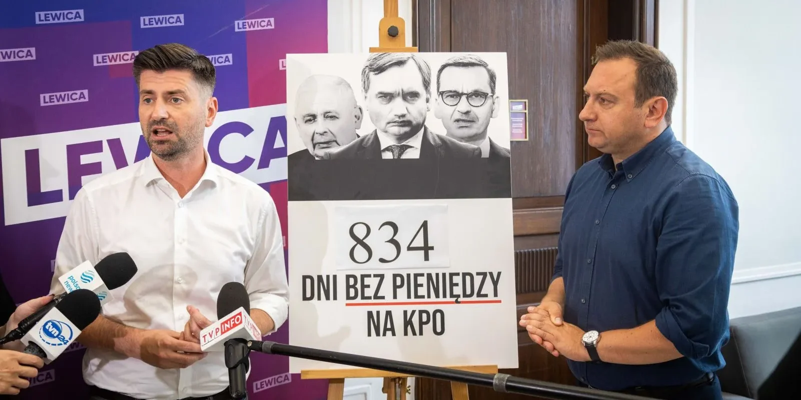 Dwóch mężczyzn podczas konferencji prasowej Lewicy, stojących obok plakatu z napisem „834 dni bez pieniędzy na KPO” i wizerunkami czterech osób, przy mikrofonach, na tle baneru z logotypem Lewica.