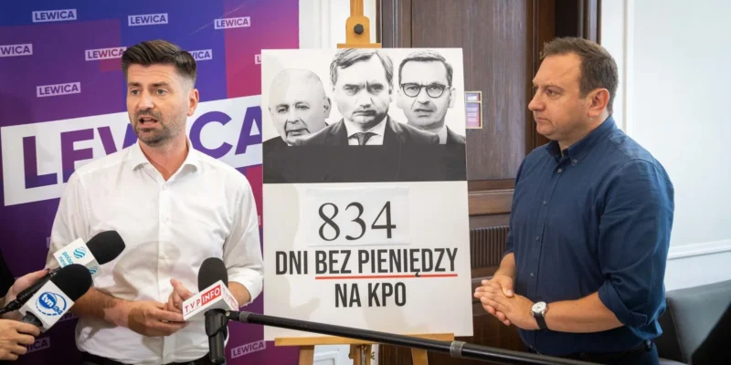 Dwóch mężczyzn podczas konferencji prasowej Lewicy, stojących obok plakatu z napisem „834 dni bez pieniędzy na KPO” i wizerunkami czterech osób, przy mikrofonach, na tle baneru z logotypem Lewica.