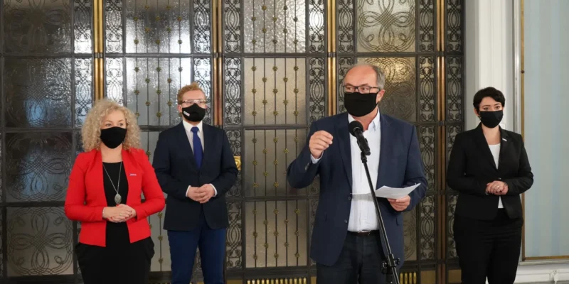 Czterech polityków w maseczkach stoi przy mikrofonie podczas konferencji prasowej, na tle ozdobnych metalowych drzwi.