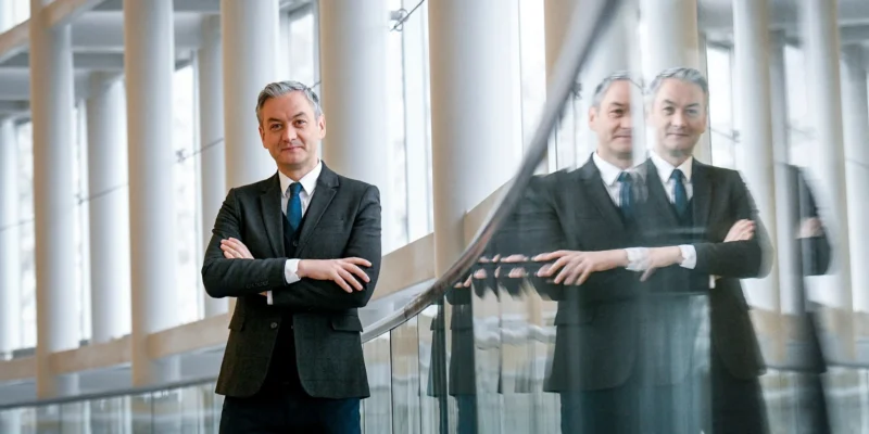 Na zdjęciu stoi elegancko ubrany mężczyzna z założonymi rękoma, a w tle widać jego odbicie w szklanej balustradzie i nowoczesnym wnętrzu.