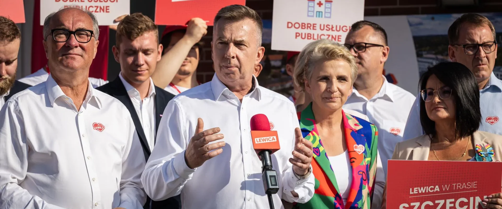 Grupa polityków Lewicy podczas konferencji prasowej, z mężczyzną w białej koszuli mówiącym do mikrofonu, na tle plakatów z logiem Lewicy i hasłem „Dobre usługi publiczne”.