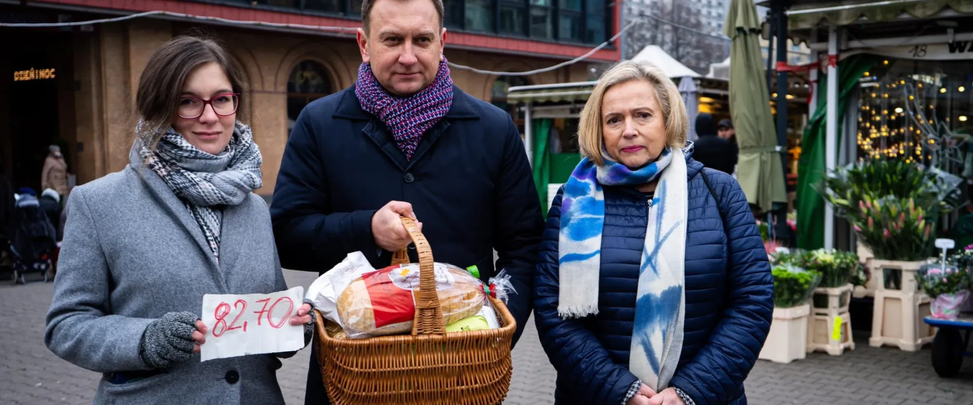 Trzy osoby stoją na targu miejskim: z lewej kobieta trzyma kartkę z ceną 82,70, po środku mężczyzna trzyma kosz z pieczywem i zakupami, a z prawej stoi kobieta w niebieskiej kurtce.