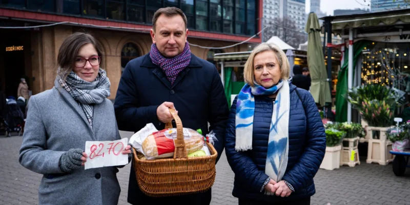 Trzy osoby stoją na targu miejskim: z lewej kobieta trzyma kartkę z ceną 82,70, po środku mężczyzna trzyma kosz z pieczywem i zakupami, a z prawej stoi kobieta w niebieskiej kurtce.