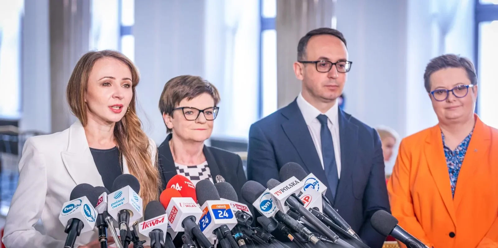 Kobieta w białym garniturze przemawia do mikrofonów podczas konferencji prasowej, towarzyszą jej trzej współpracownicy.