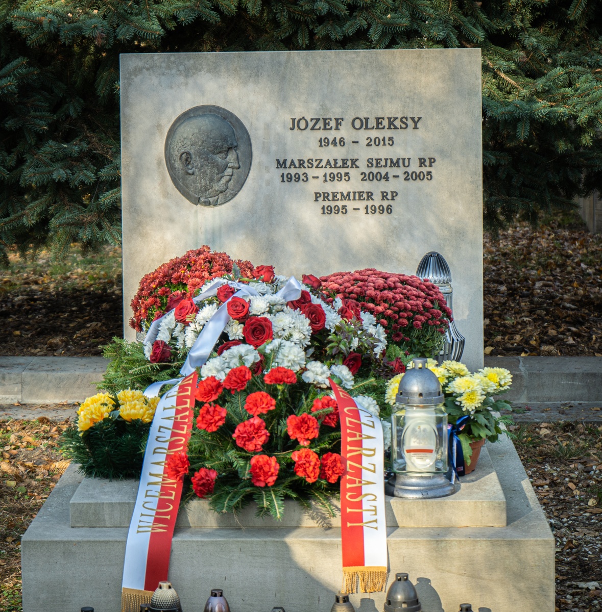 Nagrobek z medalionem przedstawiającym profil mężczyzny, otoczony kolorowymi kwiatami, wieńcami i lampionami, na tle iglastych drzew.