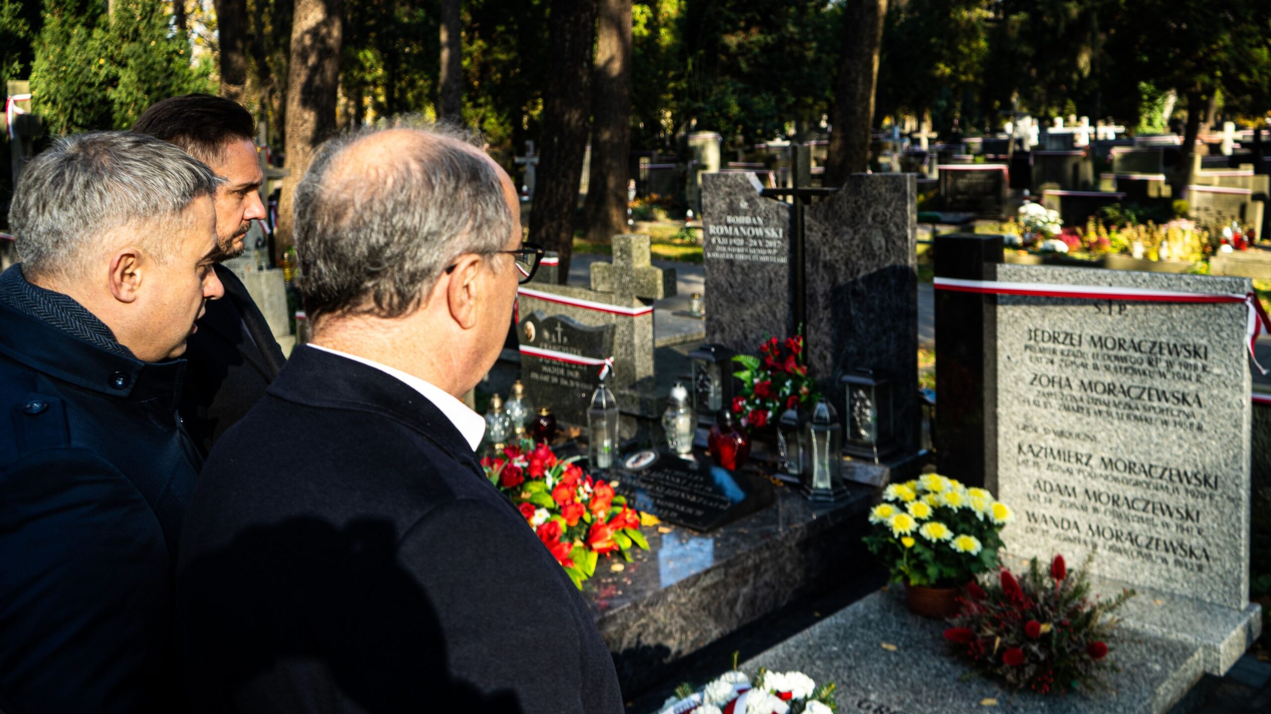 Trzej mężczyźni stoją przy grobie na cmentarzu, otoczeni nagrobkami, kwiatami i zniczami.