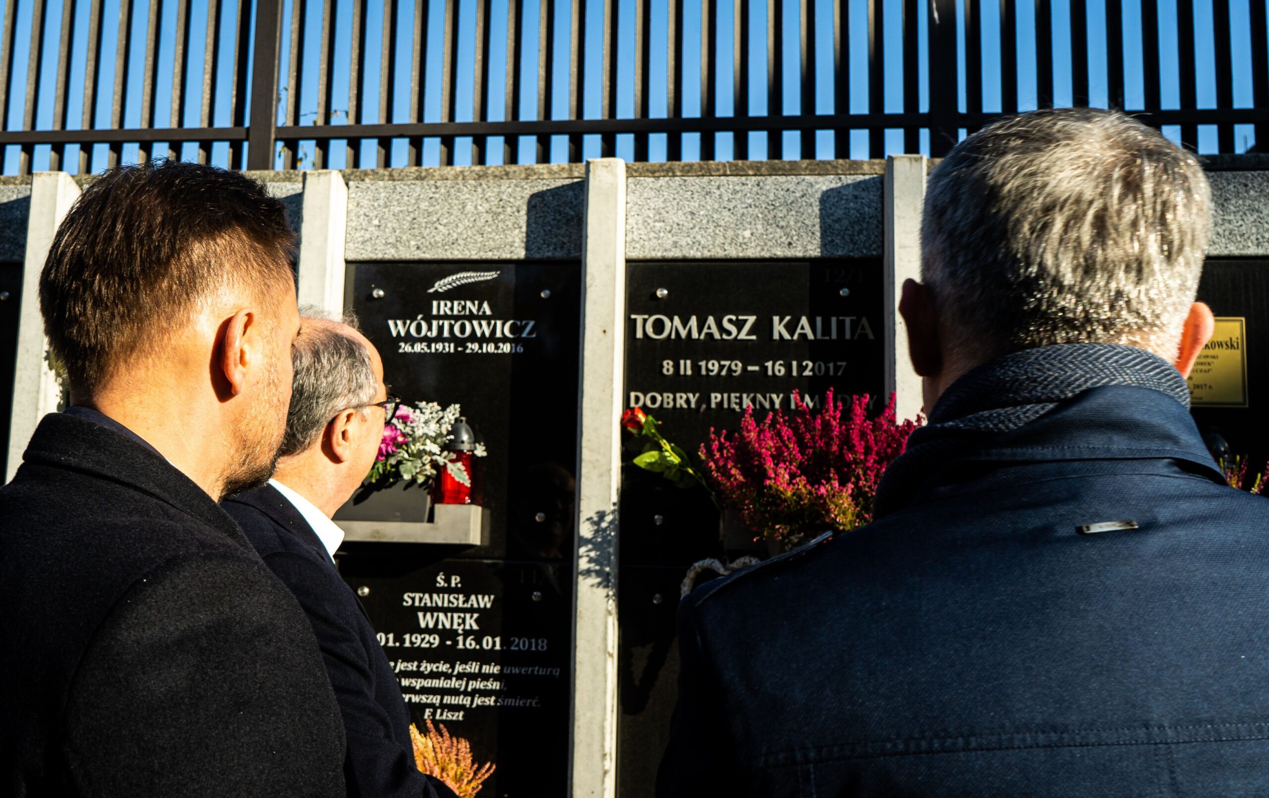 Trzy osoby widziane od tyłu stoją przed nagrobkami z kwiatami na cmentarzu, na grobach widnieją imiona i daty.