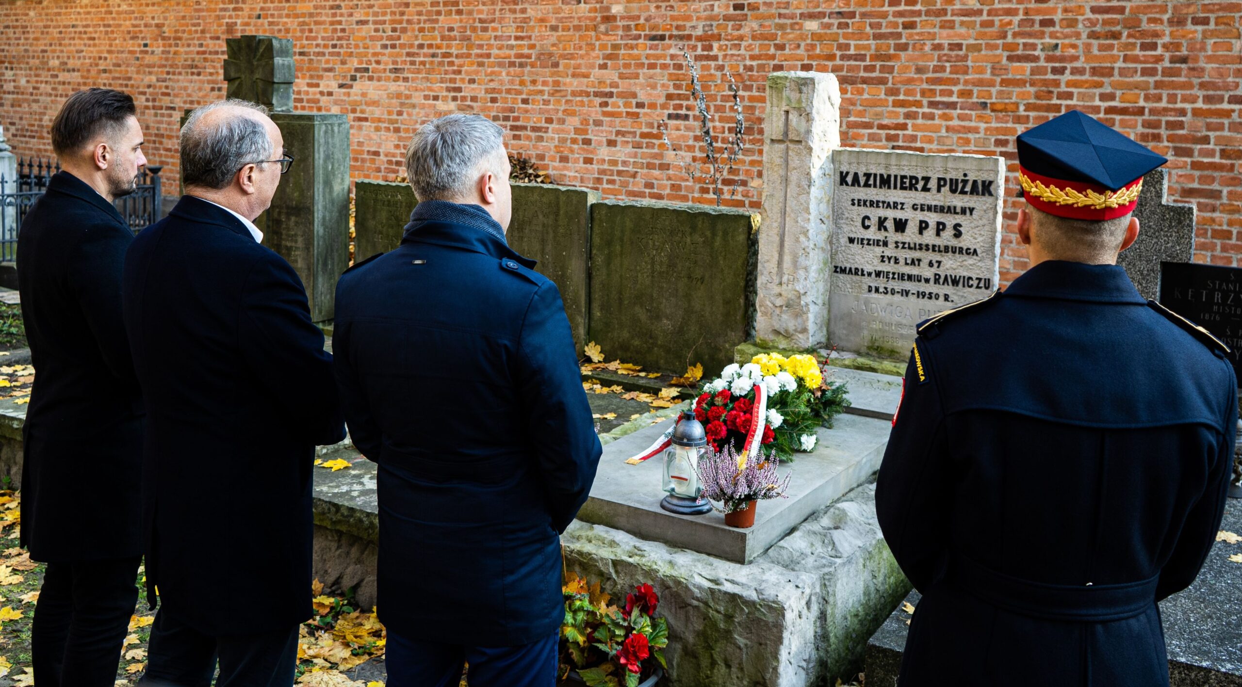 Czterej mężczyźni w czarnych płaszczach oraz mundurze wojskowym stoją w ciszy przed grobem Kazimierza Pużaka, składając hołd pamięci zmarłego.