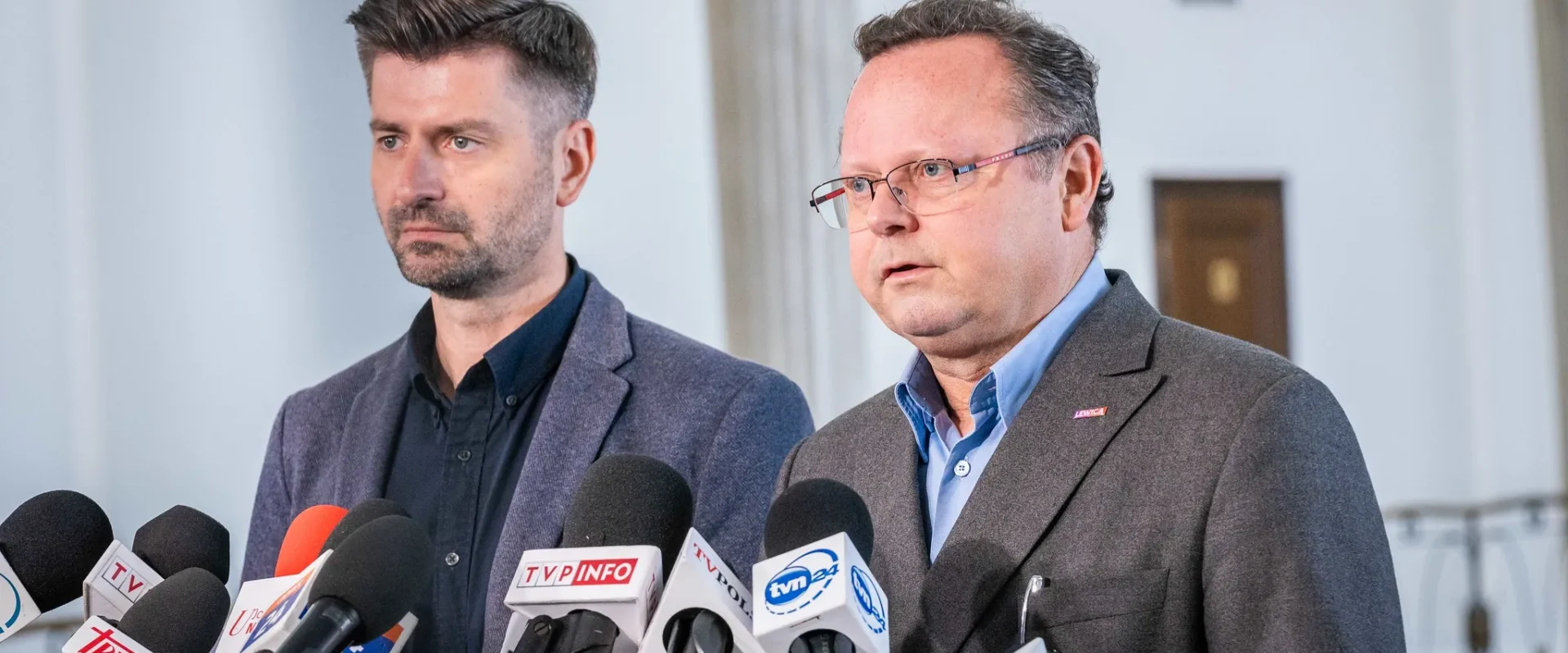 Dwóch mężczyzn w garniturach stoi przy konferencji prasowej, otoczeni zestawem mikrofonów z logotypami mediów.