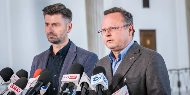 Dwóch mężczyzn w garniturach stoi przy konferencji prasowej, otoczeni zestawem mikrofonów z logotypami mediów.