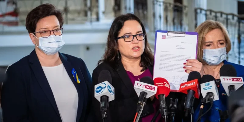 Trzy kobiety w maseczkach stojące przed mikrofonami podczas konferencji prasowej; centrę trzyma kartę z dokumentem, a na ubraniu jednej z nich widnieje niebiesko‑żółta wstążka wspierająca Ukrainę.