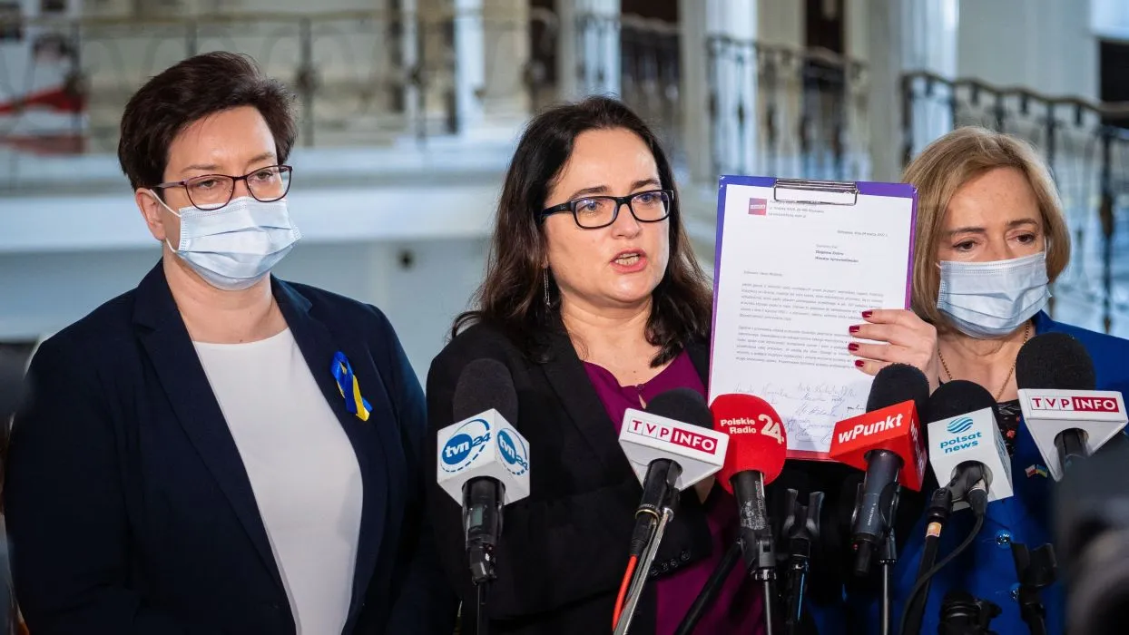 Trzy kobiety w maseczkach stojące przed mikrofonami podczas konferencji prasowej; centrę trzyma kartę z dokumentem, a na ubraniu jednej z nich widnieje niebiesko‑żółta wstążka wspierająca Ukrainę.