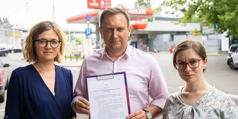 Trzy osoby – dwie kobiety i mężczyzna – stoją na tle stacji benzynowej, trzymając dokument.