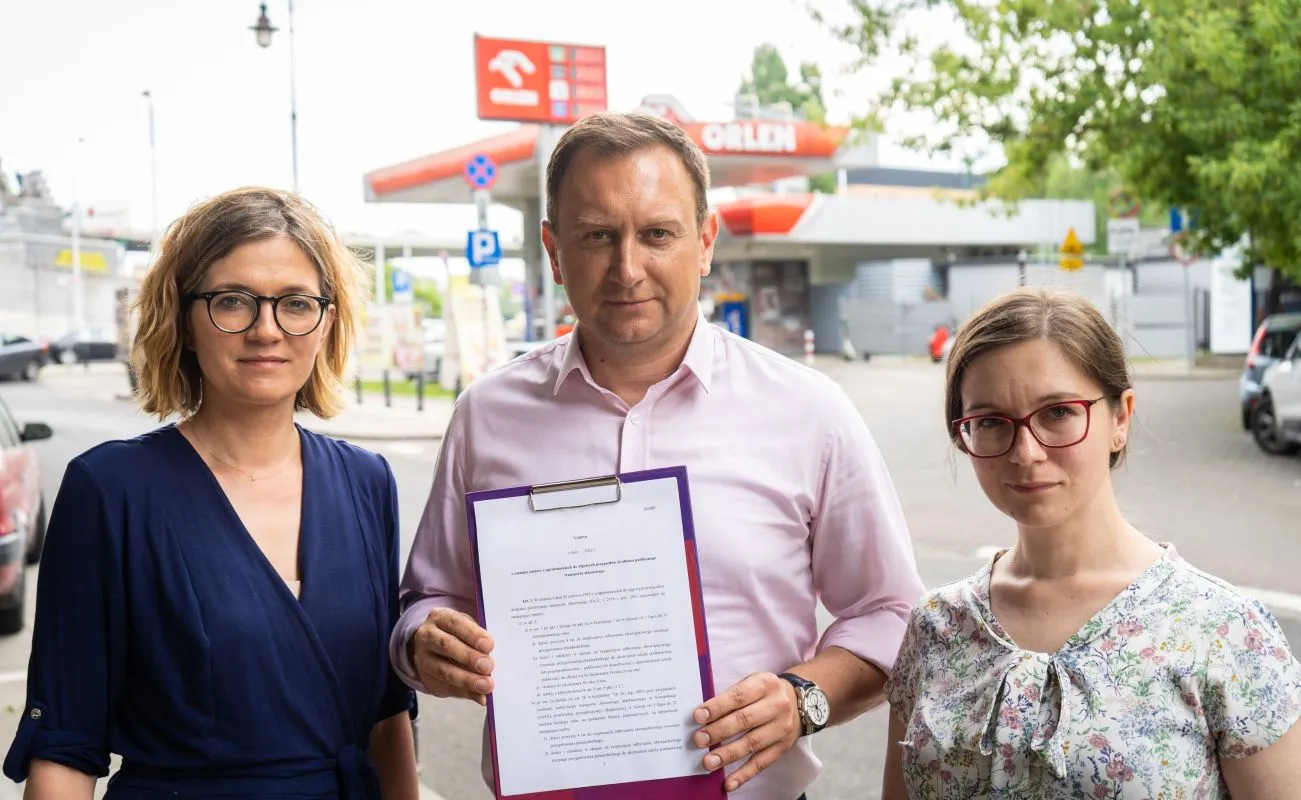 Trzy osoby – dwie kobiety i mężczyzna – stoją na tle stacji benzynowej, trzymając dokument.