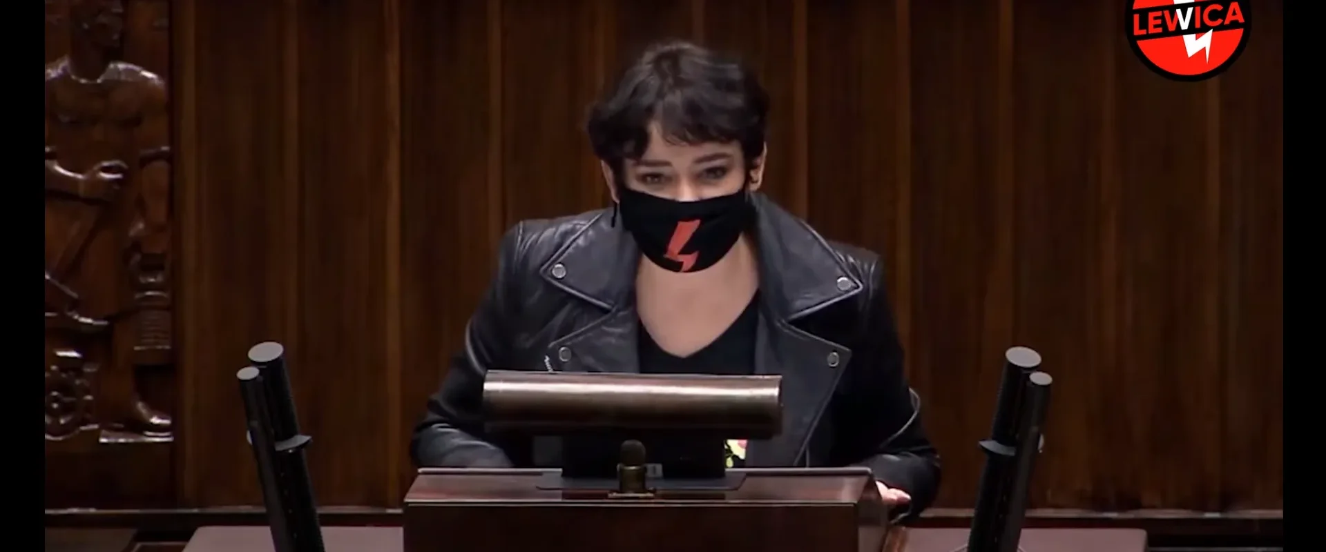 Osoba przemawia z mównicy w sali z drewnianymi panelami, ubrana w czarną skórzaną kurtkę i czarną maseczkę z czerwonym symbolem; w tle widoczny logotyp Lewica.