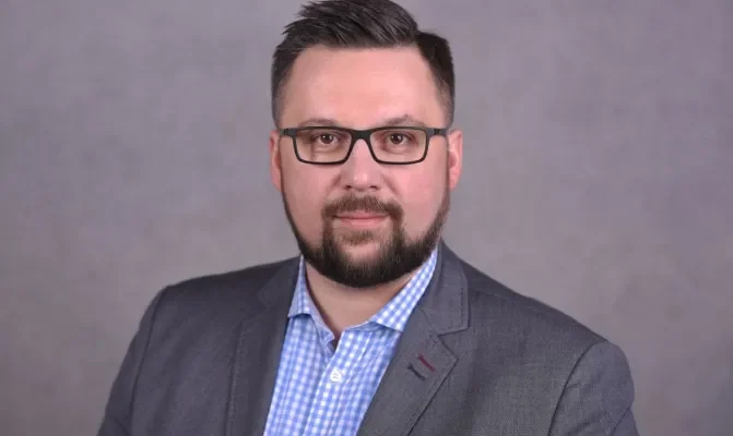 Portret biznesowy mężczyzny w szarym garniturze, okularach i koszuli w kratę na neutralnym tle.