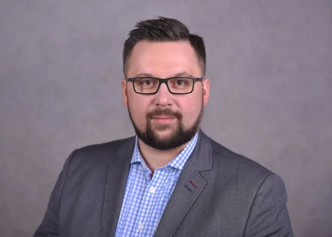 Portret biznesowy mężczyzny w szarym garniturze, okularach i koszuli w kratę na neutralnym tle.