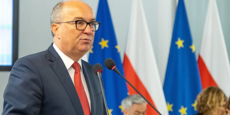 Mężczyzna w garniturze mówi do mikrofonu na tle flag europejskich i polskich podczas konferencji.