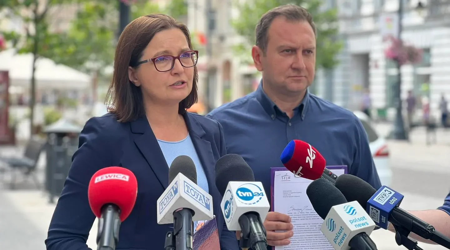 Dwoje polityków na konferencji prasowej na zewnątrz, kobieta w okularach i granatowej marynarce przemawia do mikrofonów, obok niej stoi mężczyzna, a w tle widać miejską ulicę z kwitnącymi drzewami.