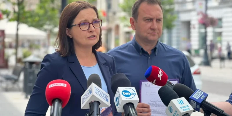 Dwoje polityków na konferencji prasowej na zewnątrz, kobieta w okularach i granatowej marynarce przemawia do mikrofonów, obok niej stoi mężczyzna, a w tle widać miejską ulicę z kwitnącymi drzewami.