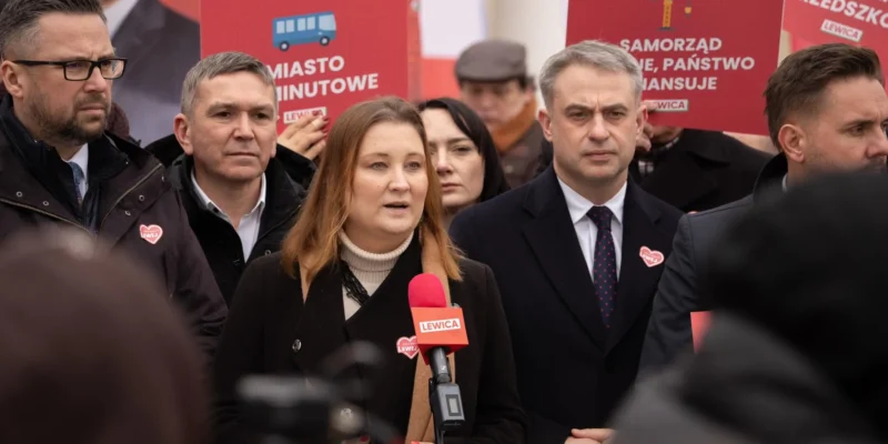 Kobieta przemawia do mikrofonu podczas wiecu Lewicy, otoczona przez mężczyzn i czerwone plakaty z logo Lewicy w tle.