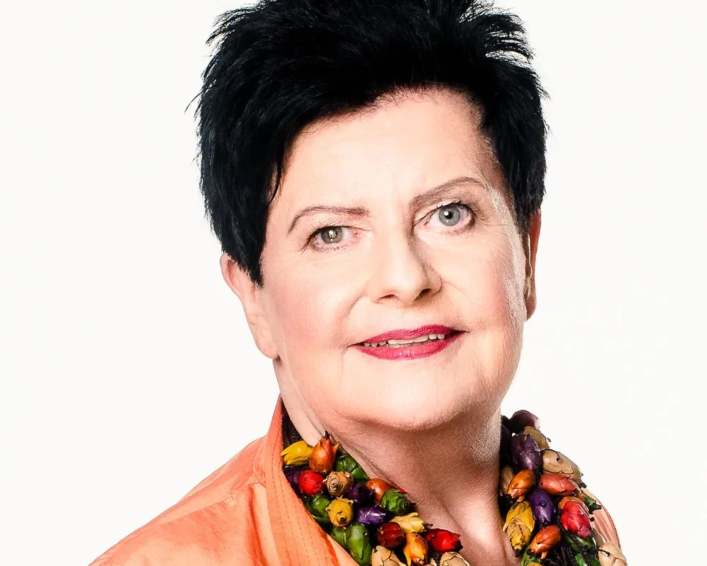 Portret kobiety z krótkimi czarnymi włosami, uśmiechniętej, w pomarańczowej bluzce i kolorowym naszyjniku z koralików na jasnym tle.