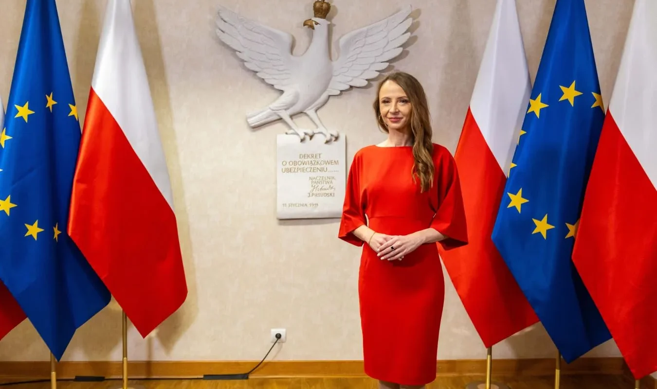 Kobieta w czerwonej sukience stoi między flagami Unii Europejskiej i Polski, na tle ściany z białym orłem i tablicą upamiętniającą.