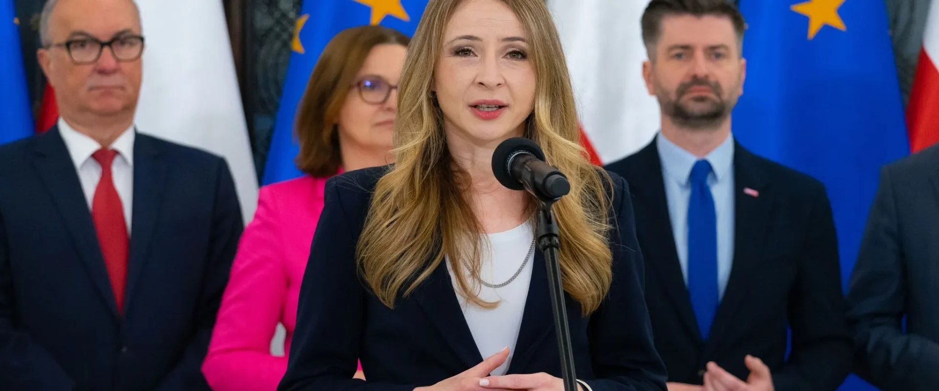 Kobieta w granatowym garniturze przemawia do mikrofonu podczas konferencji prasowej, stojąc na tle flag Unii Europejskiej i innych polityków w garniturach.