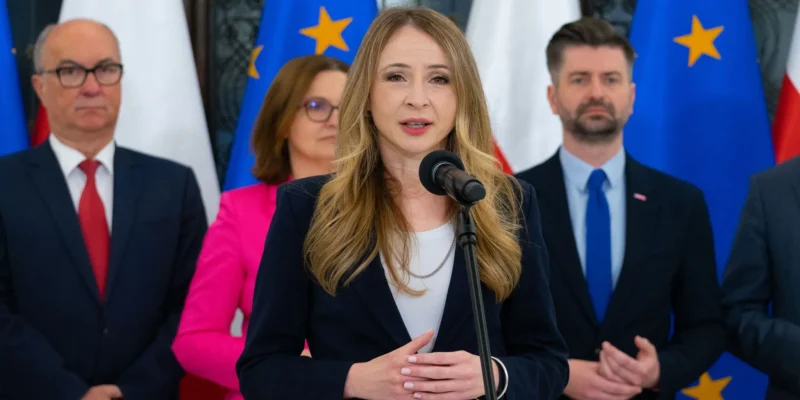 Kobieta w granatowym garniturze przemawia do mikrofonu podczas konferencji prasowej, stojąc na tle flag Unii Europejskiej i innych polityków w garniturach.