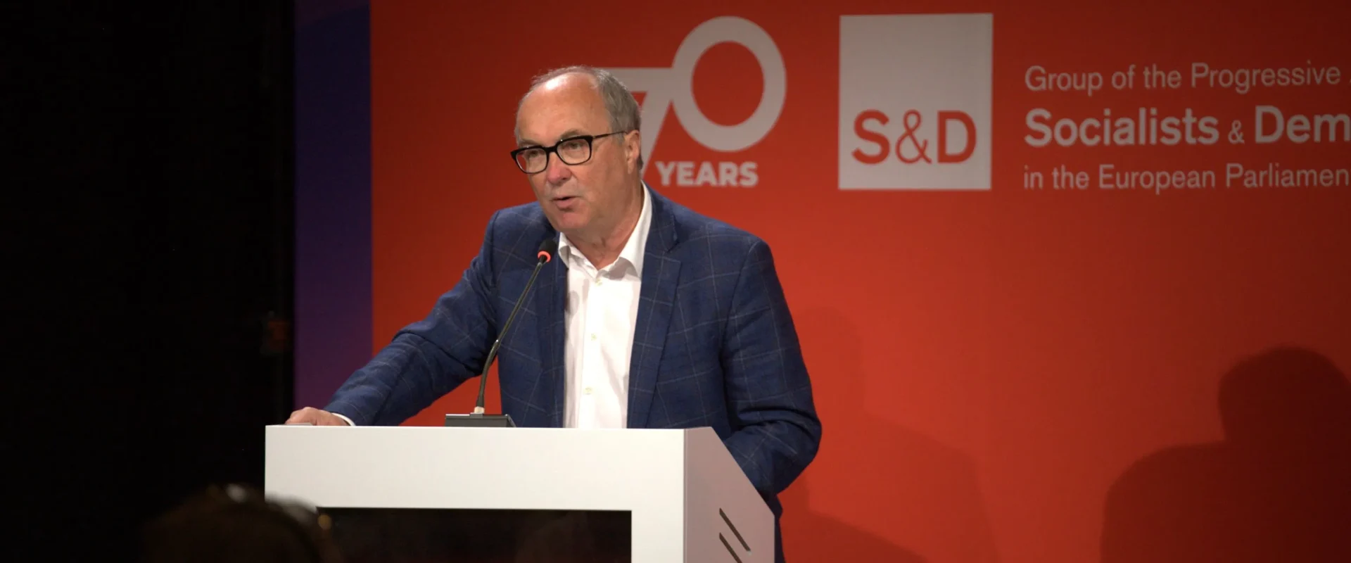 Mężczyzna w okularach przemawia z mównicy, na tle czerwonej planszy z logo S&D z okazji 70-lecia.