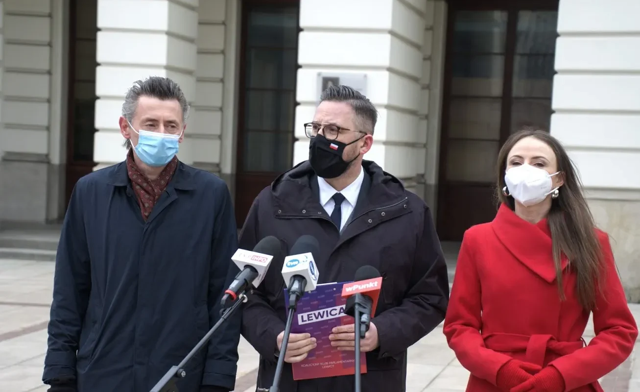 Trzy osoby w maseczkach stojące przed mikrofonami na zewnątrz budynku podczas konferencji prasowej, a w centrum mężczyzna trzyma plakat z napisem Lewica w kontekście Narodowego Programu Kopernikańskiego.