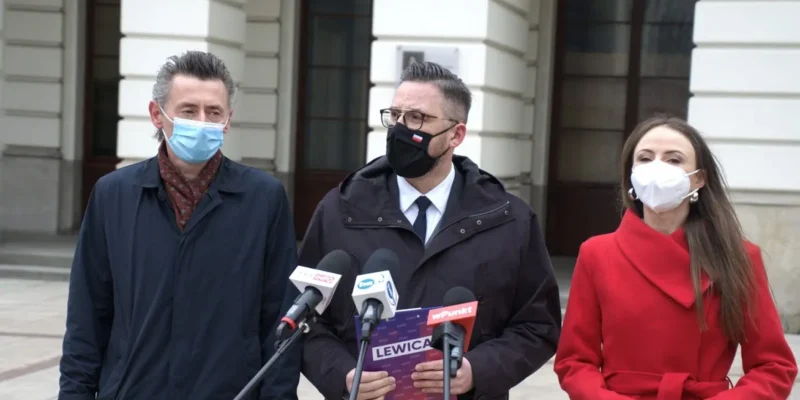 Trzy osoby w maseczkach stojące przed mikrofonami na zewnątrz budynku podczas konferencji prasowej, a w centrum mężczyzna trzyma plakat z napisem Lewica w kontekście Narodowego Programu Kopernikańskiego.
