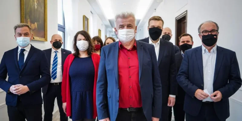 Grupa polityków Lewicy w formalnych garniturach i maseczkach stoi w jasnym korytarzu; na pierwszym planie mężczyzna w granatowej marynarce i czerwonej koszuli, kandydat Lewicy na Rzecznika Praw Obywatelskich.