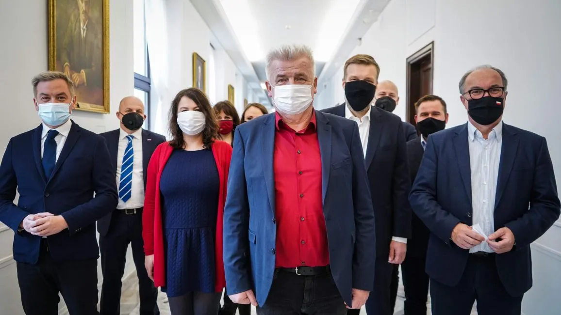 Grupa polityków Lewicy w formalnych garniturach i maseczkach stoi w jasnym korytarzu; na pierwszym planie mężczyzna w granatowej marynarce i czerwonej koszuli, kandydat Lewicy na Rzecznika Praw Obywatelskich.