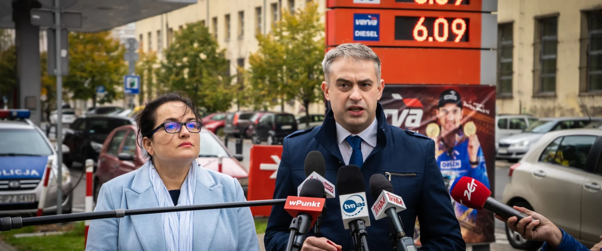 Dwóch polityków podczas konferencji prasowej, stojących przy mikrofonach, na tle tablicy z cenami paliw 6.09.