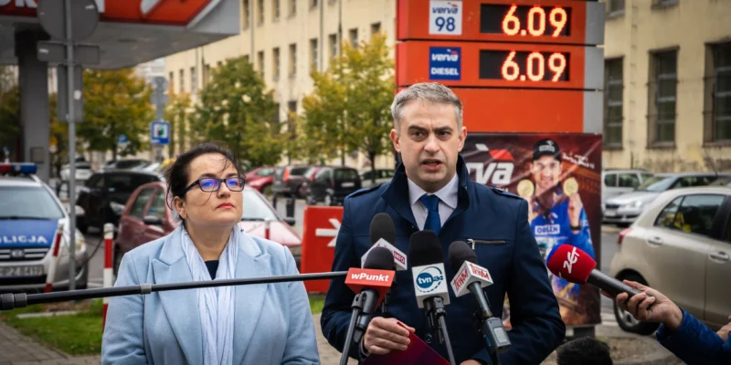 Dwóch polityków podczas konferencji prasowej, stojących przy mikrofonach, na tle tablicy z cenami paliw 6.09.