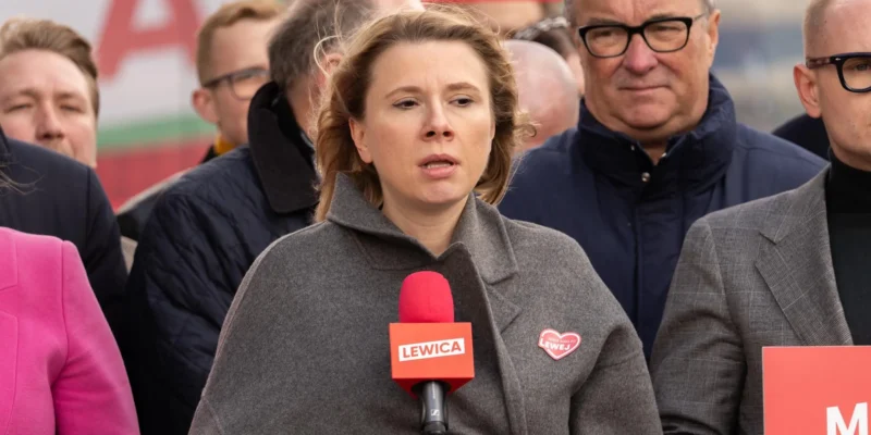 Kobieta przemawia do mikrofonu podczas konferencji, w otoczeniu innych osób; na czerwonym mikrofonie widnieje napis Lewica.