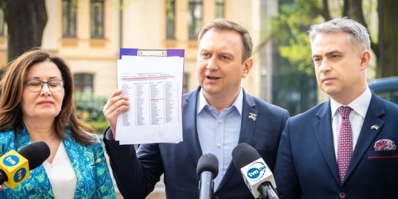 Na zdjęciu trzech polityków uczestniczy w konferencji prasowej, podczas której jeden z nich trzyma dokument z wyraźnie widoczną listą, a obok znajdują się mikrofony mediów.