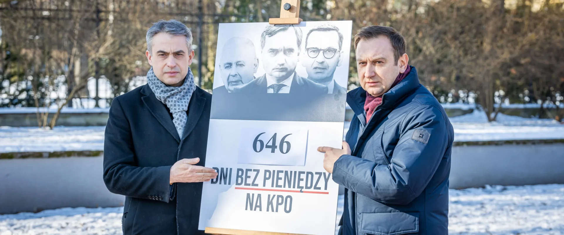 Dwóch mężczyzn stoi w zimnym parku i prezentuje plakat na sztaludze z czarno-białymi wizerunkami polityków oraz napisem 646 dni bez pieniędzy na KPO.