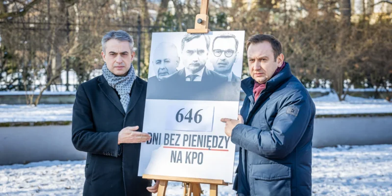 Dwóch mężczyzn stoi w zimnym parku i prezentuje plakat na sztaludze z czarno-białymi wizerunkami polityków oraz napisem 646 dni bez pieniędzy na KPO.