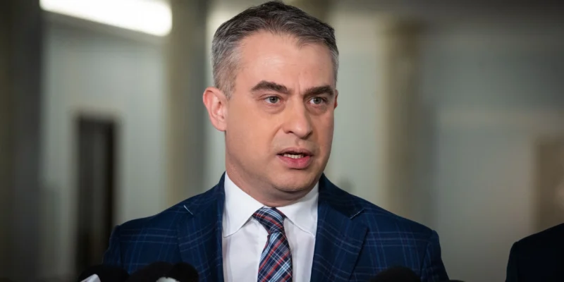 Polityk w granatowym garniturze w kratę, białej koszuli i kraciastym krawacie przemawia do mikrofonów podczas konferencji prasowej.