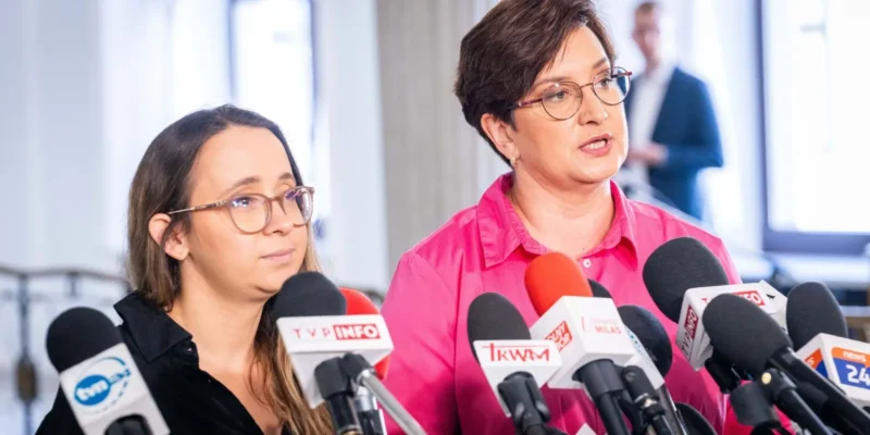 Dwie kobiety podczas konferencji prasowej stoją za licznymi mikrofonami i udzielają wypowiedzi mediom.