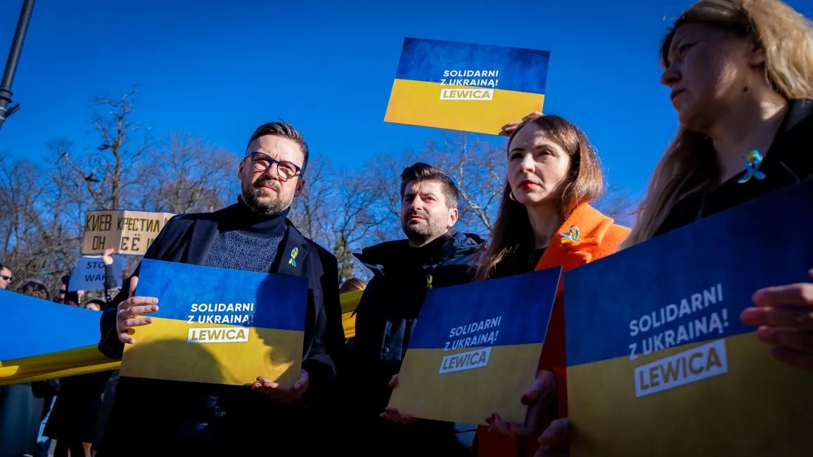 Grupa ludzi trzymających transparenty z napisem "Solidarni z Ukrainą!" na tle błękitnego nieba.