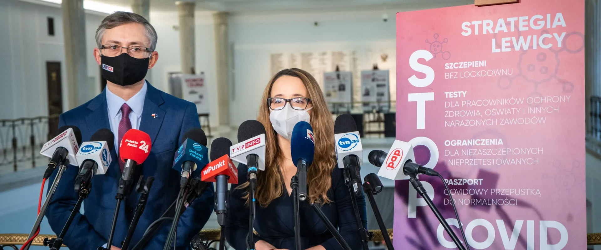 Na zdjęciu widać dwóch polityków podczas konferencji prasowej, obok nich znajdują się mikrofony oraz plakat z informacjami o strategii dotyczącej COVID-19.