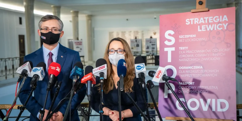 Na zdjęciu widać dwóch polityków podczas konferencji prasowej, obok nich znajdują się mikrofony oraz plakat z informacjami o strategii dotyczącej COVID-19.