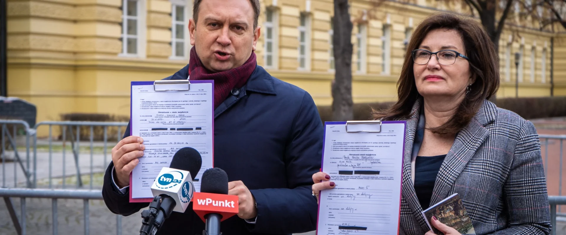 Dwóch ludzi na konferencji prasowej przed żółtym budynkiem trzyma dokumenty przypięte do clipboardów i przemawia do mediów.