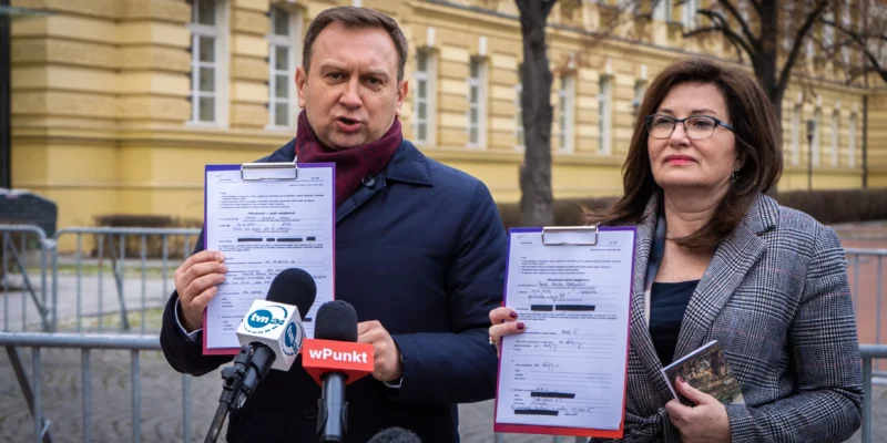 Dwóch ludzi na konferencji prasowej przed żółtym budynkiem trzyma dokumenty przypięte do clipboardów i przemawia do mediów.