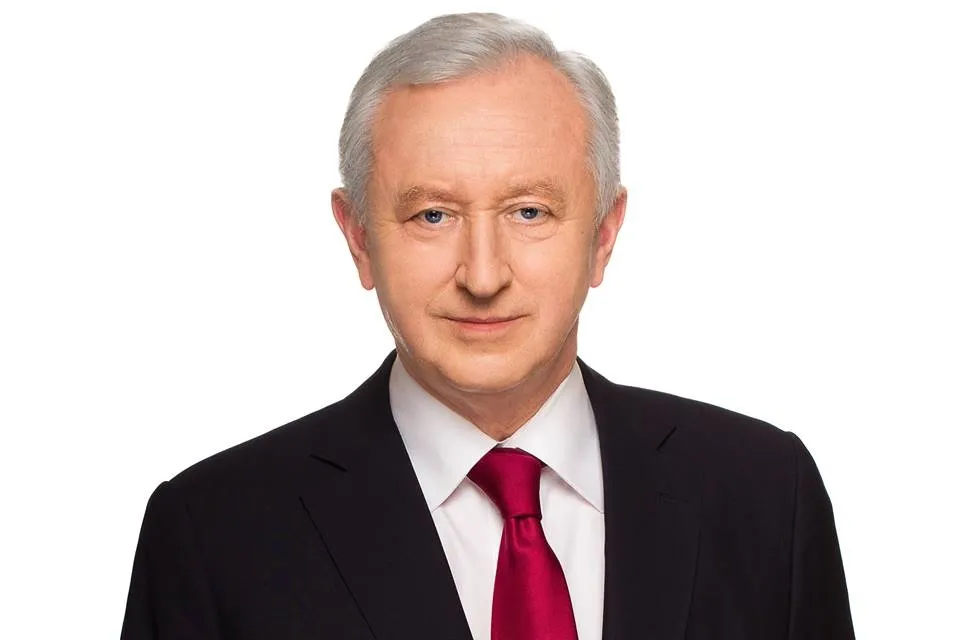 Portret siwowłosego mężczyzny w czarnym garniturze, białej koszuli i czerwonym krawacie na białym tle, patrzącego w obiektyw.